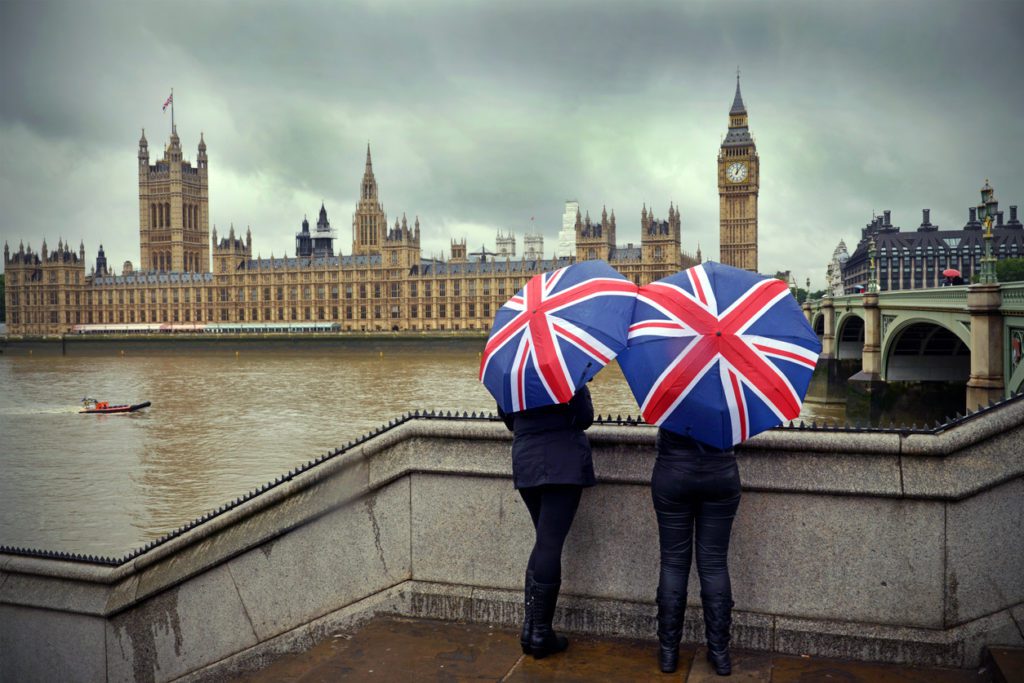 Two tourists huddle beneath British flag umbrellas showing the peculiar British weather pattern in the background